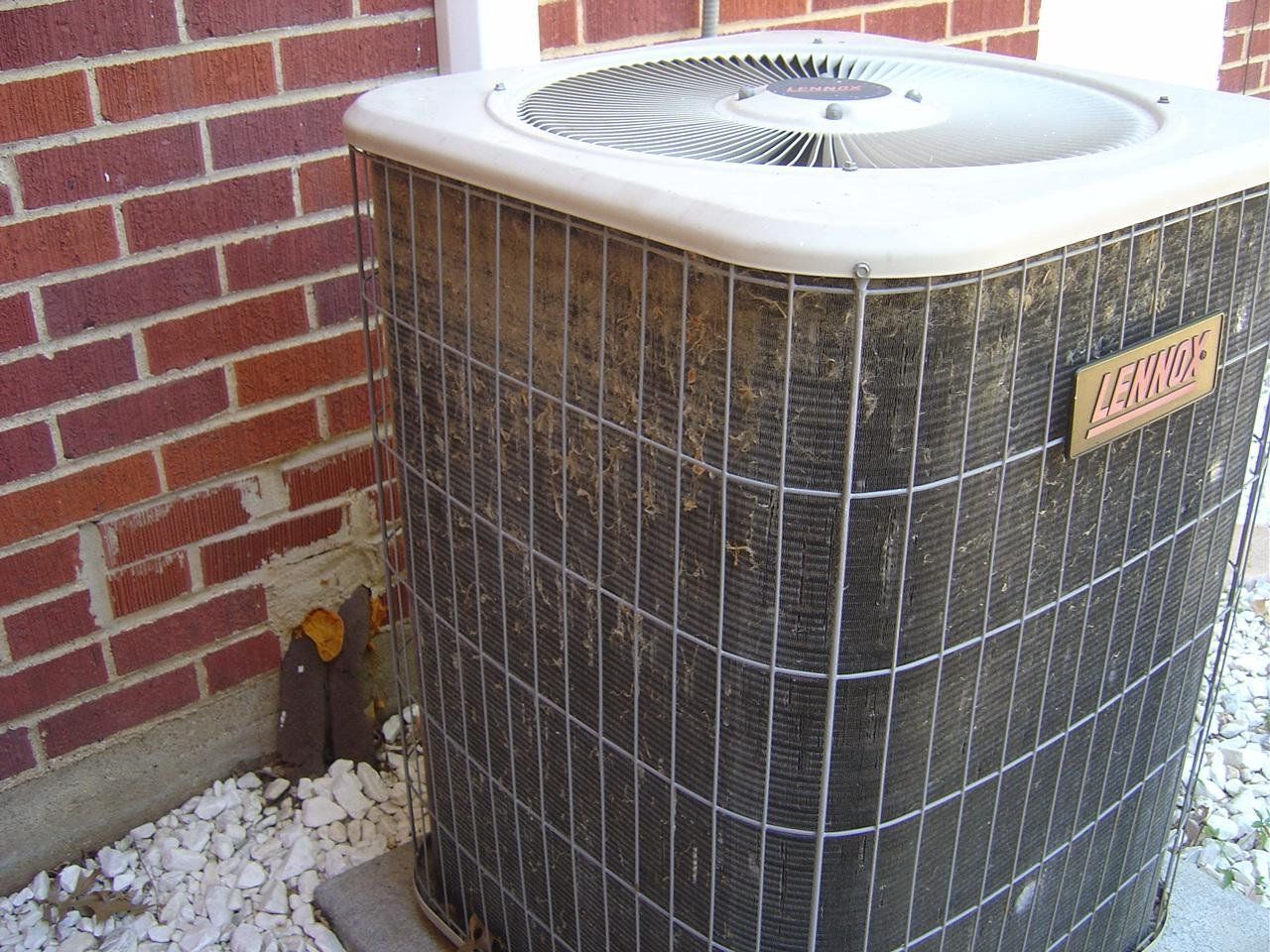 An outdoor air conditioning unit next to a brick wall. The unit is dirty with a wire cage.