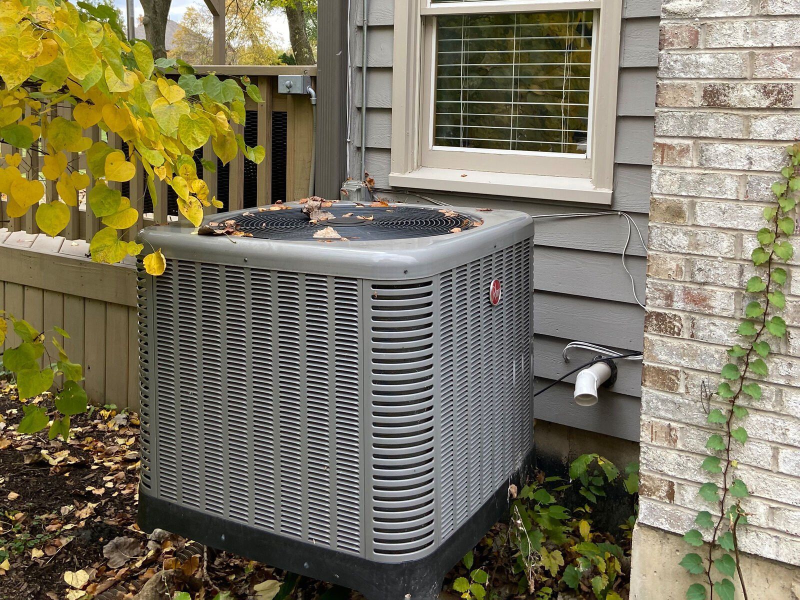 Air conditioning unit next to a house with gray siding and a brick wall.