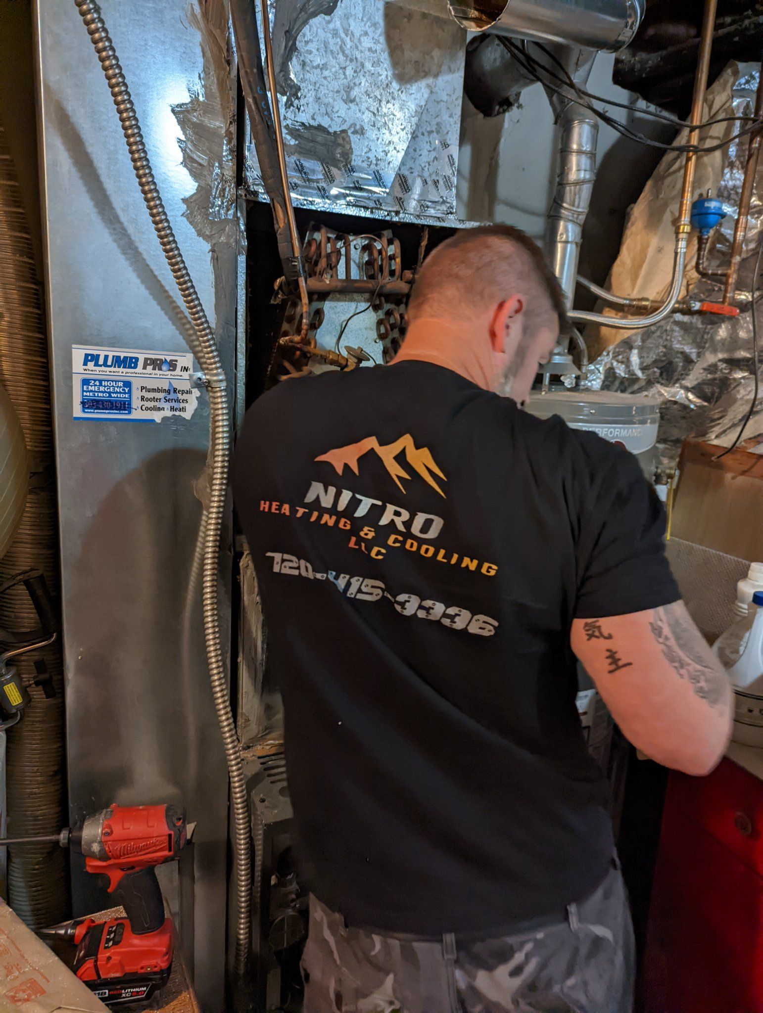 HVAC technician working on furnace, wearing a black shirt with company logo. Red drill sits nearby.