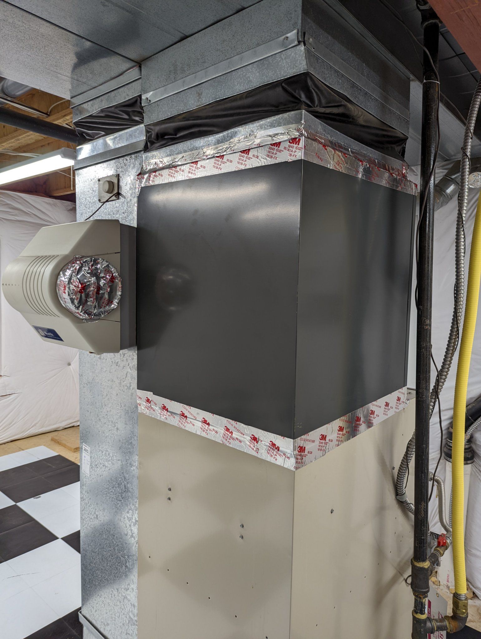 A furnace with attached humidifier in a basement setting. The unit is grey and silver with red tape.