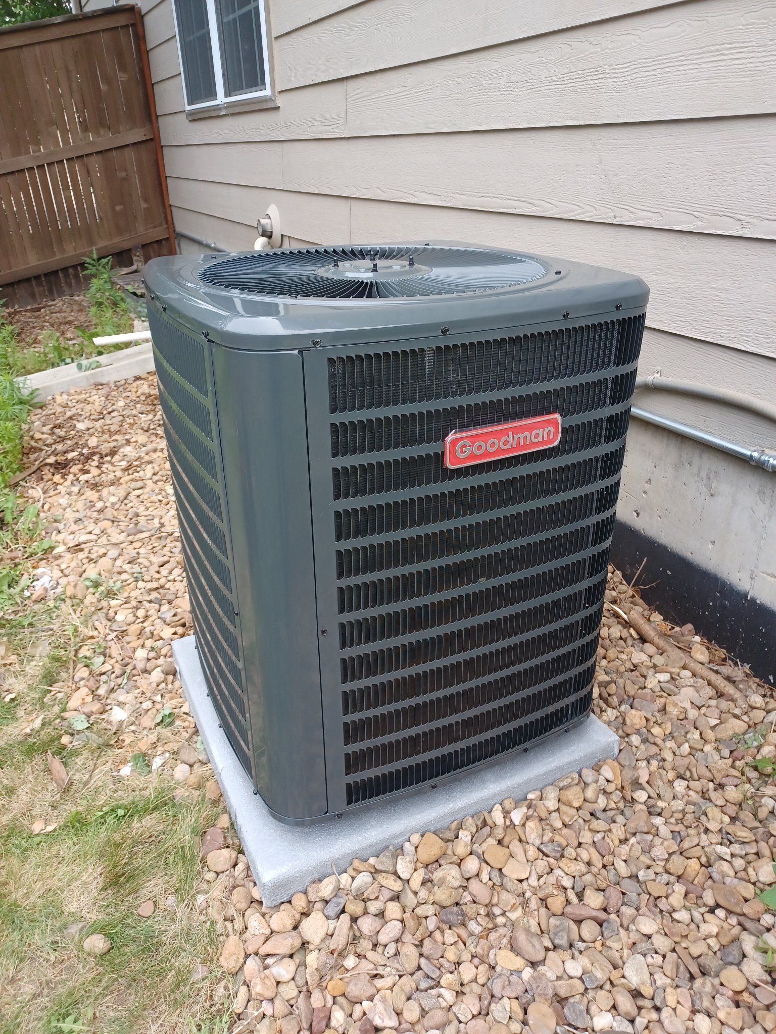 Goodman air conditioning unit on a concrete pad, surrounded by gravel and grass, next to a building.