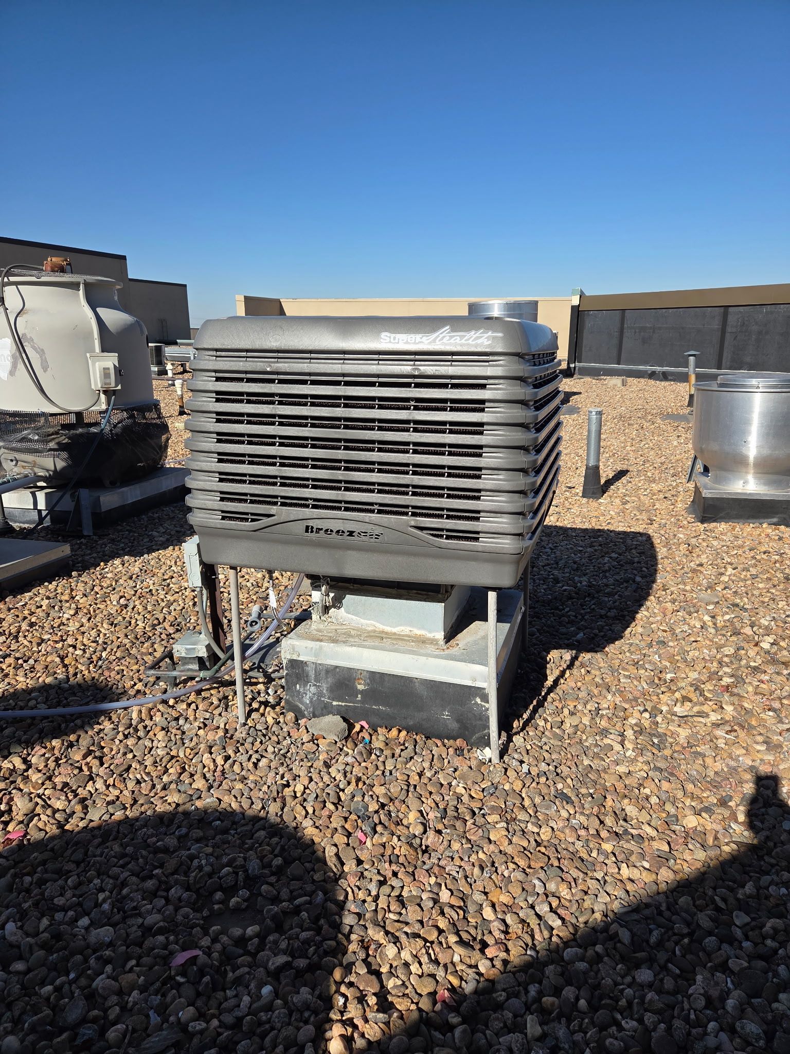 Rooftop air conditioning unit with gray metal casing and horizontal louvers on a gravel surface under a blue sky.