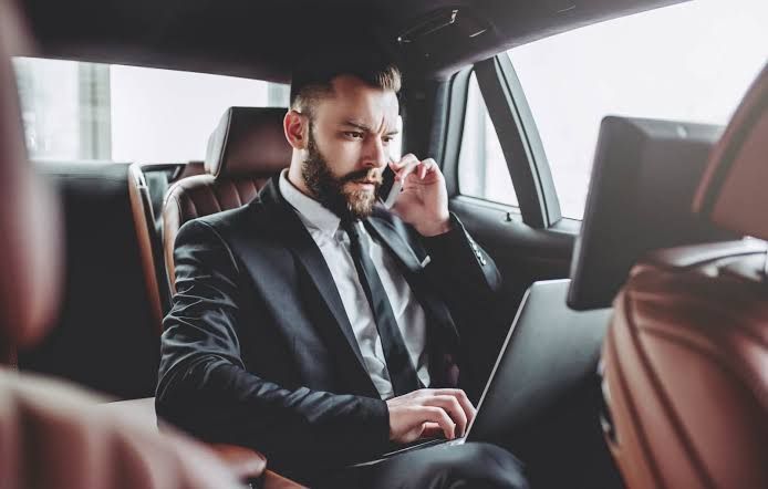 A man in a suit is sitting in the back seat of a car using a laptop and talking on a cell phone.