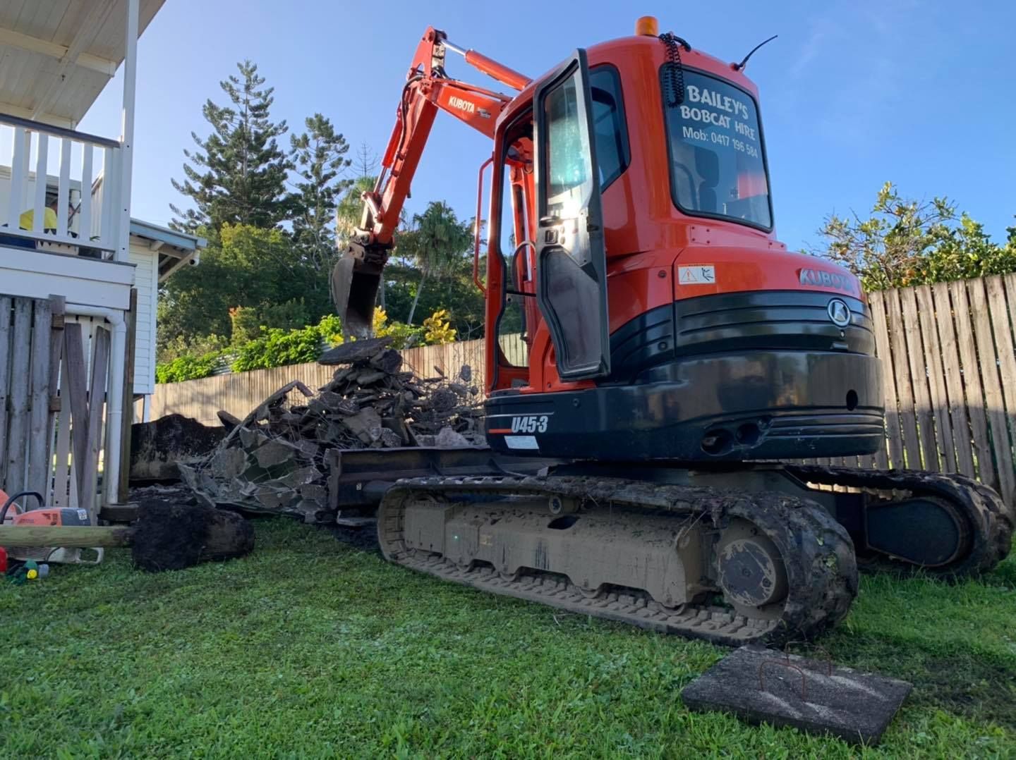 Orange Excavator Digging Dirt on A Green Lawn Near a House and Fence — Bailey's Bobcat & Excavator Hire In Dumbleton, QLD