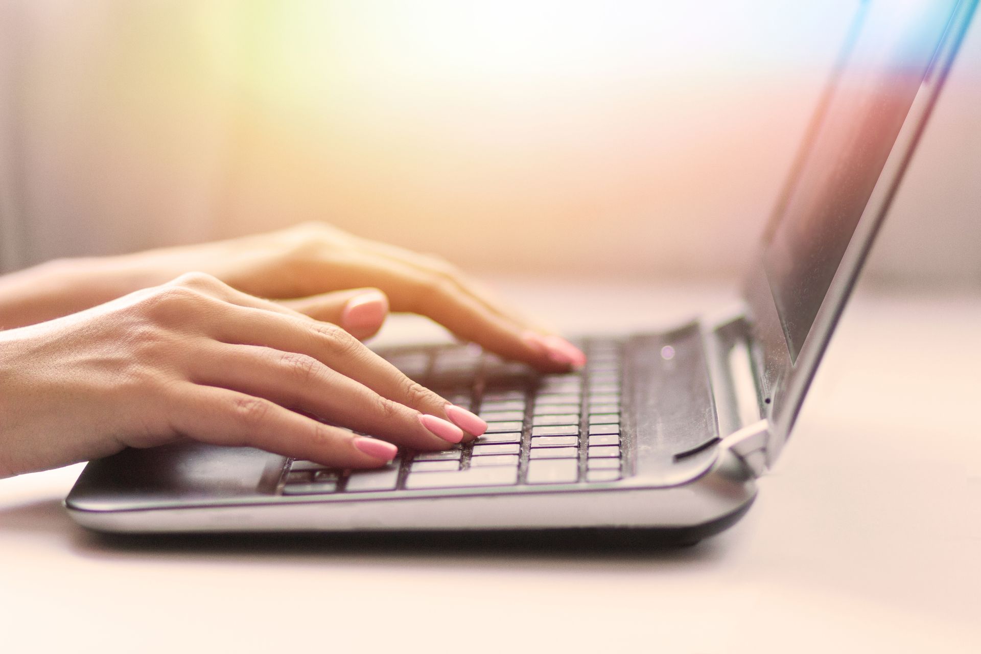 Person's hands typing on a laptop keyboard, close-up. Pink nails, bright setting.