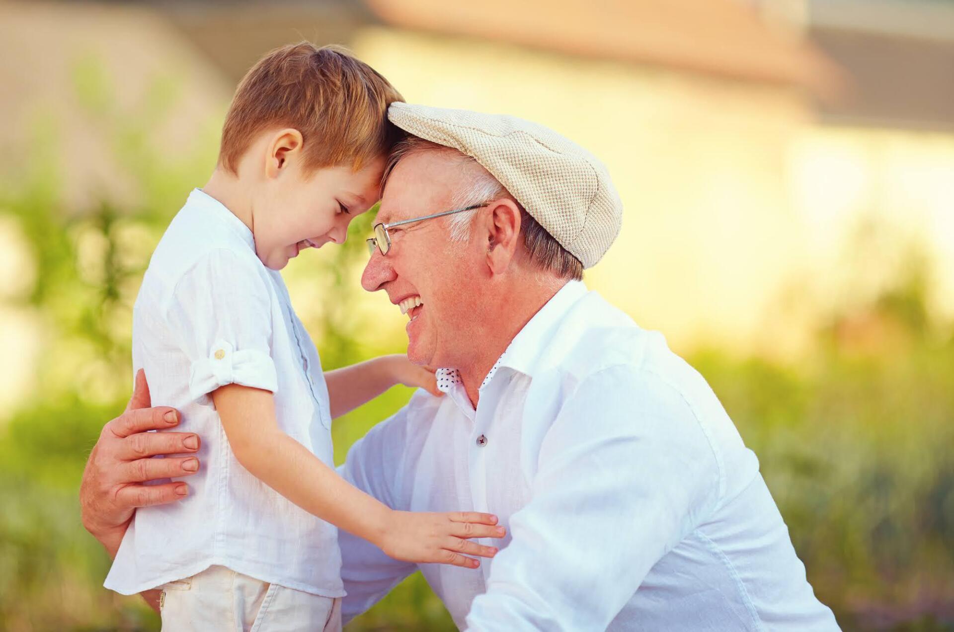 Grandfather smiles, touching foreheads with grandson in a sunny outdoor setting. Both wear white clothing.
