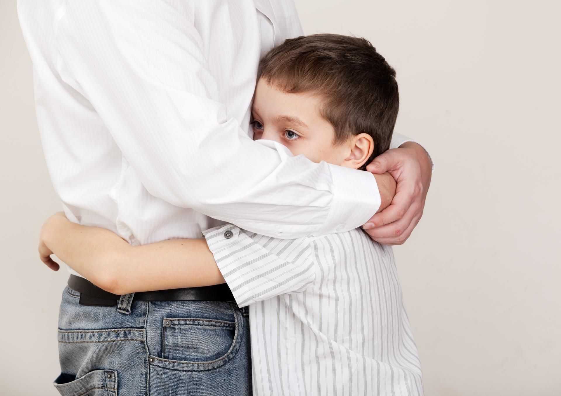 A boy hugging a man, hiding his face. Man wears white shirt and jeans. Boy wears striped shirt.