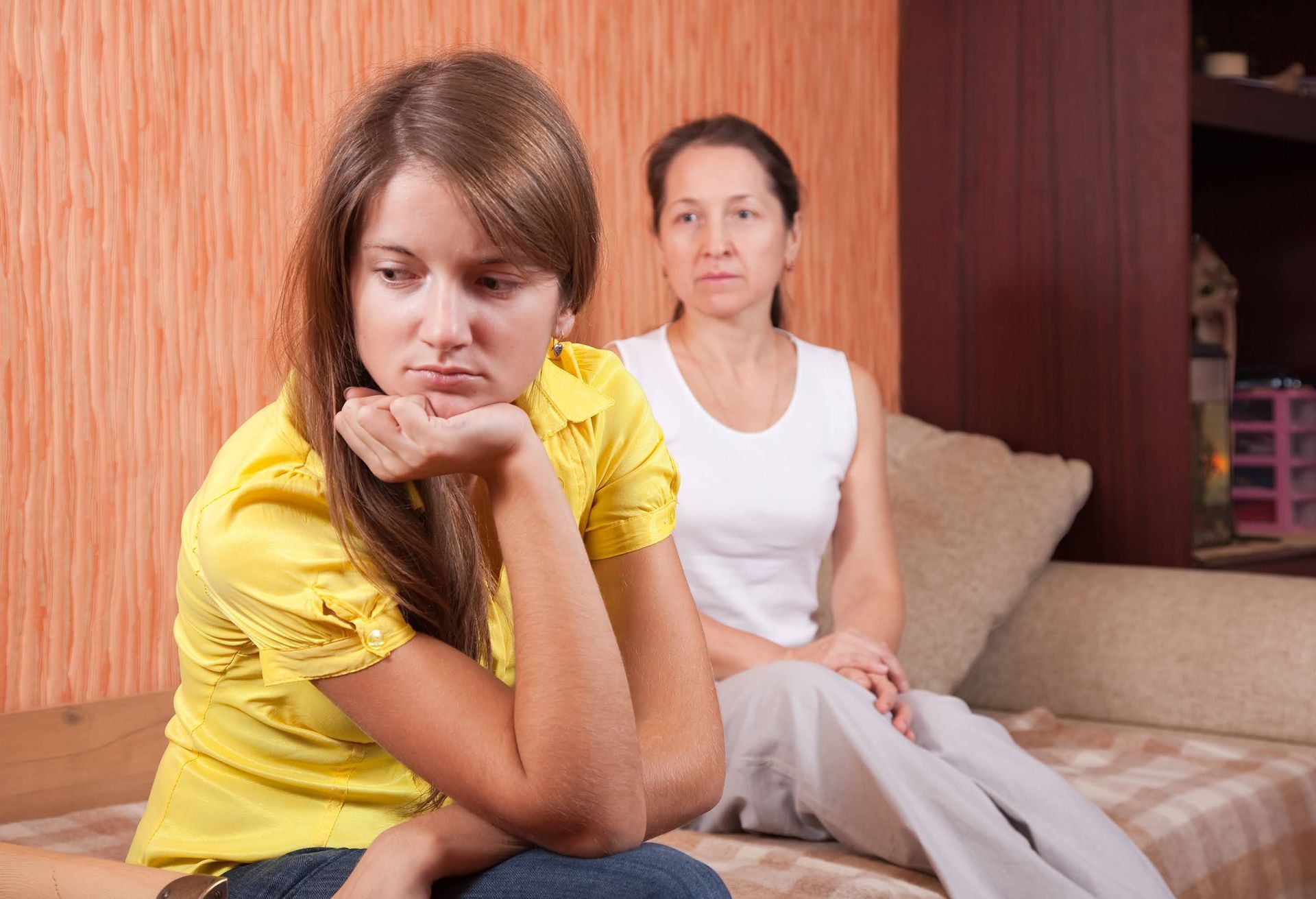 Teenager in yellow shirt looking sad, sitting next to concerned mother on a couch.