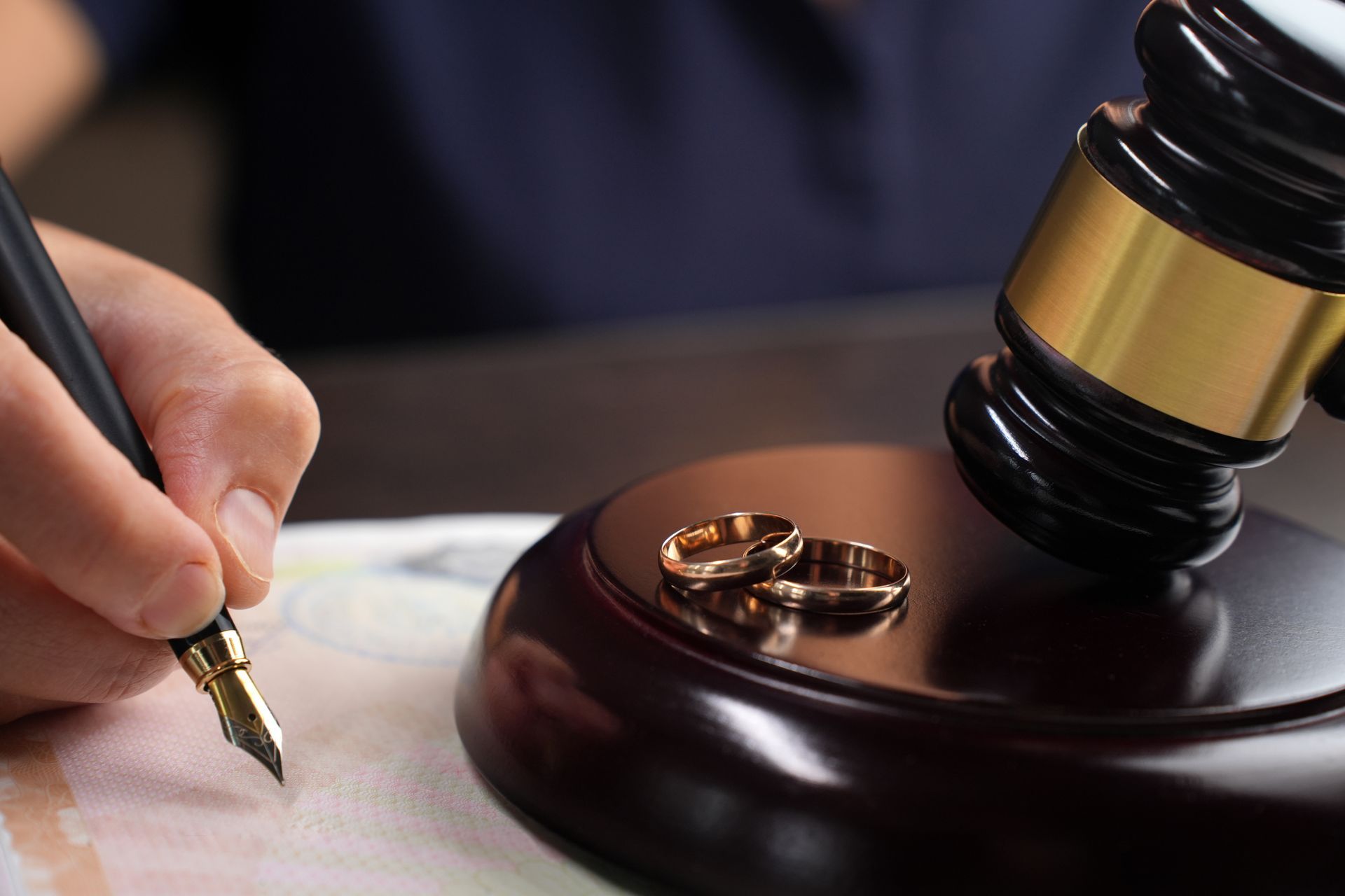 Close-up of a person signing a divorce document next to a gavel and wedding rings.