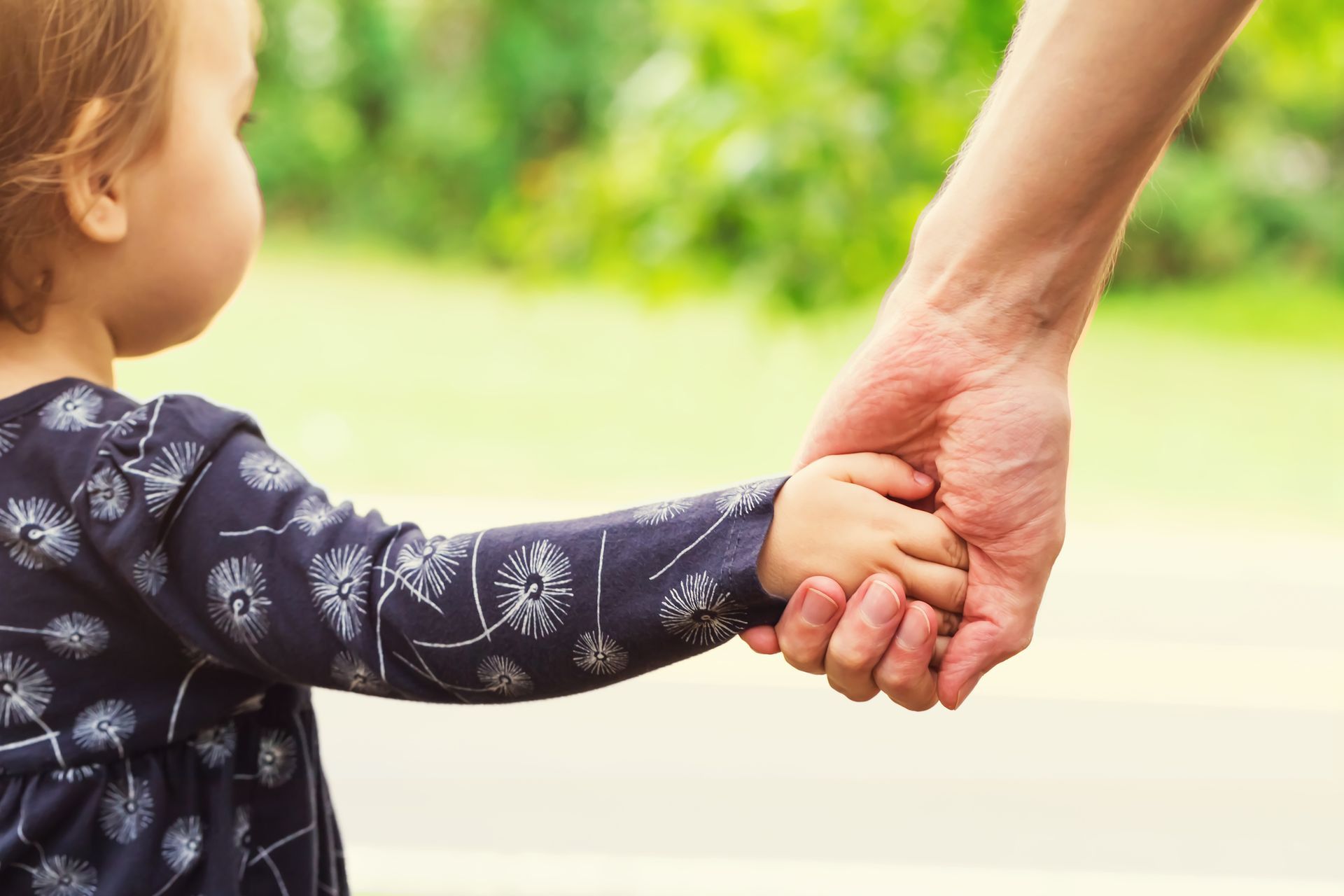 A toddler holding an adult's hand, walking outside with green foliage in the background.