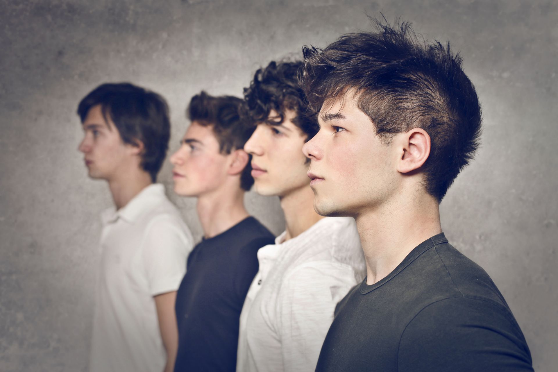 Four young men in a row, facing left. They wear different colored t-shirts against a gray wall.