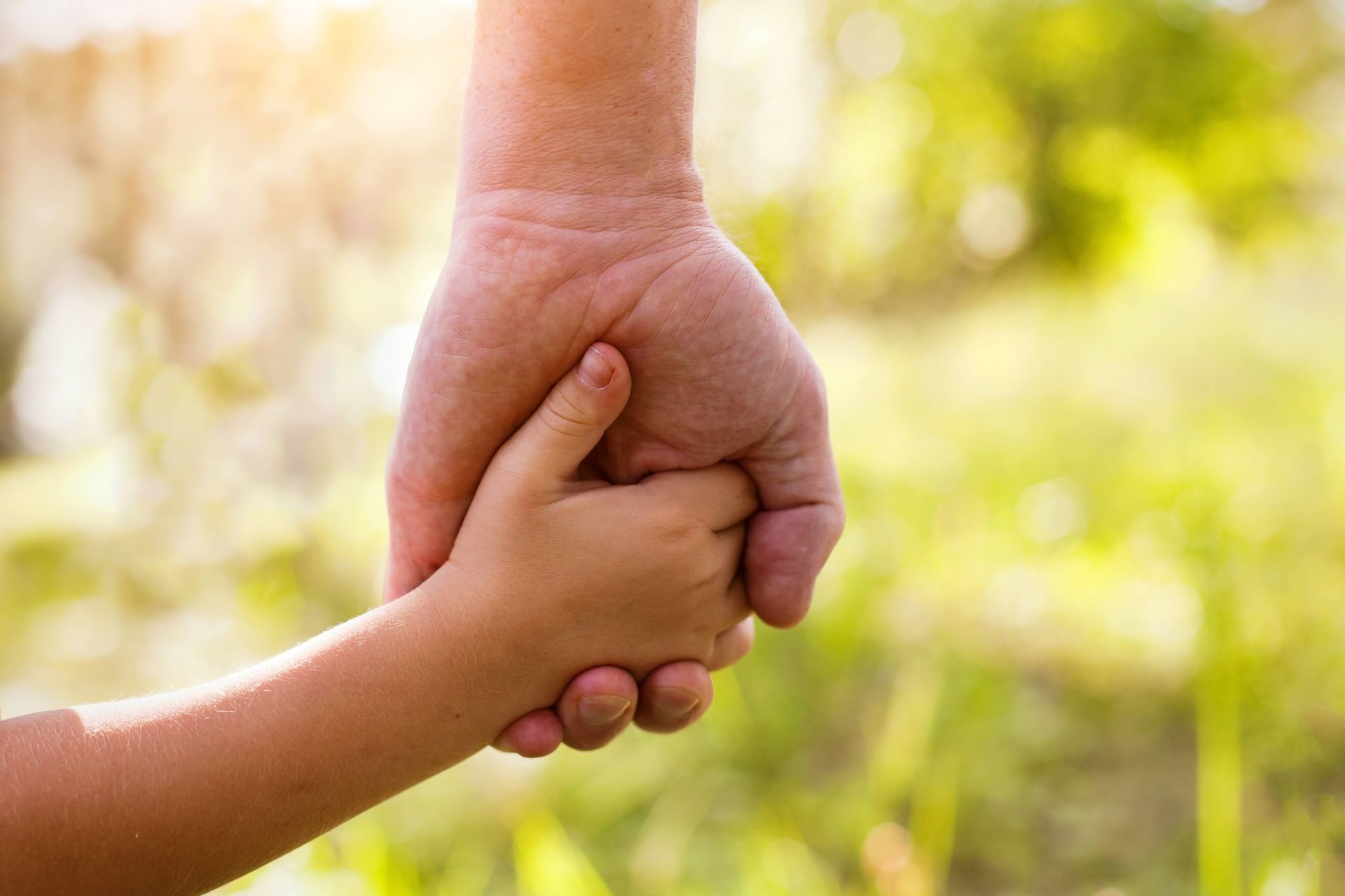 Adult hand holding a child's hand, outdoors. Focus on hands with bright sunlight.