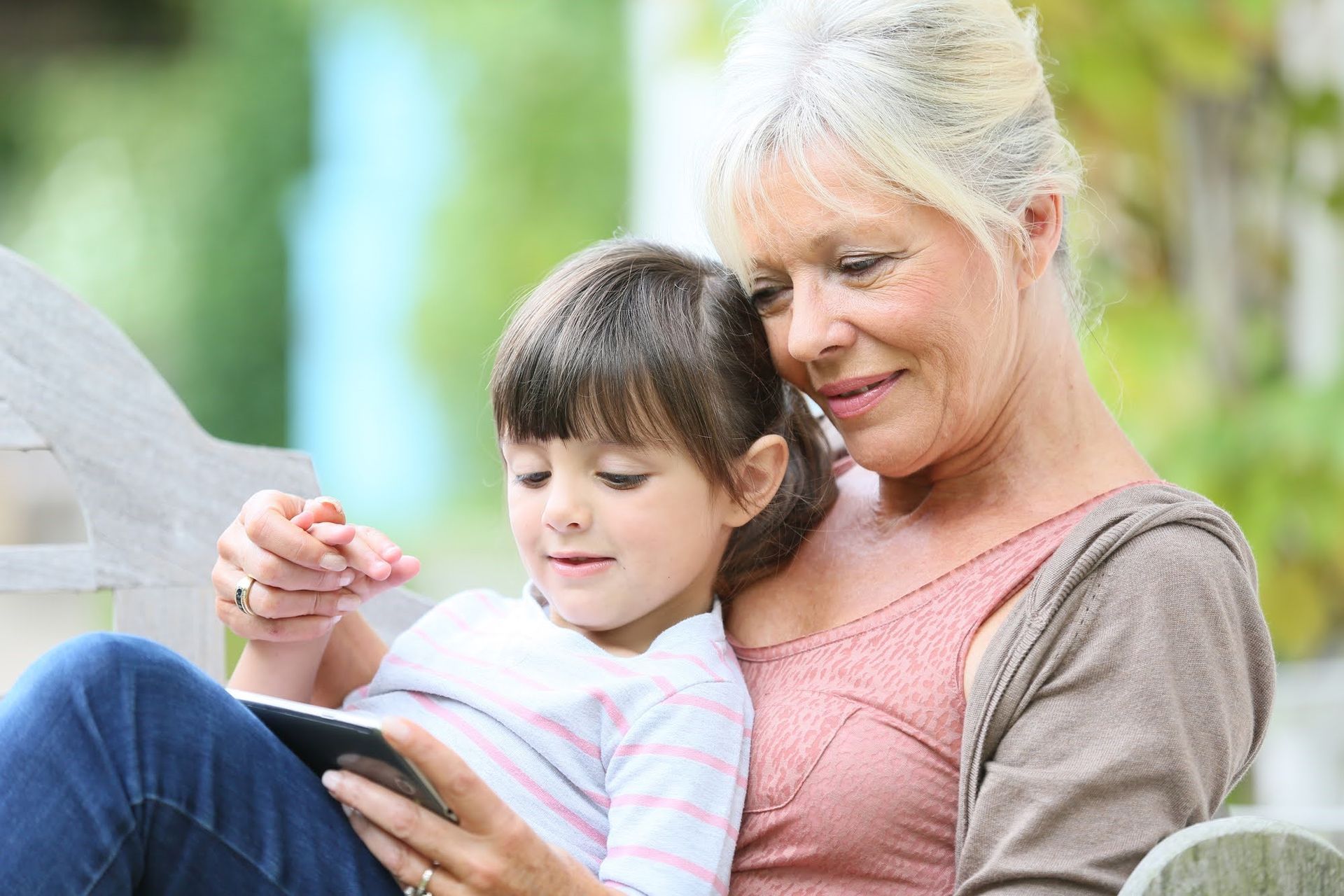 Grandmother and granddaughter sit on a bench, looking at a tablet. Outdoor setting.