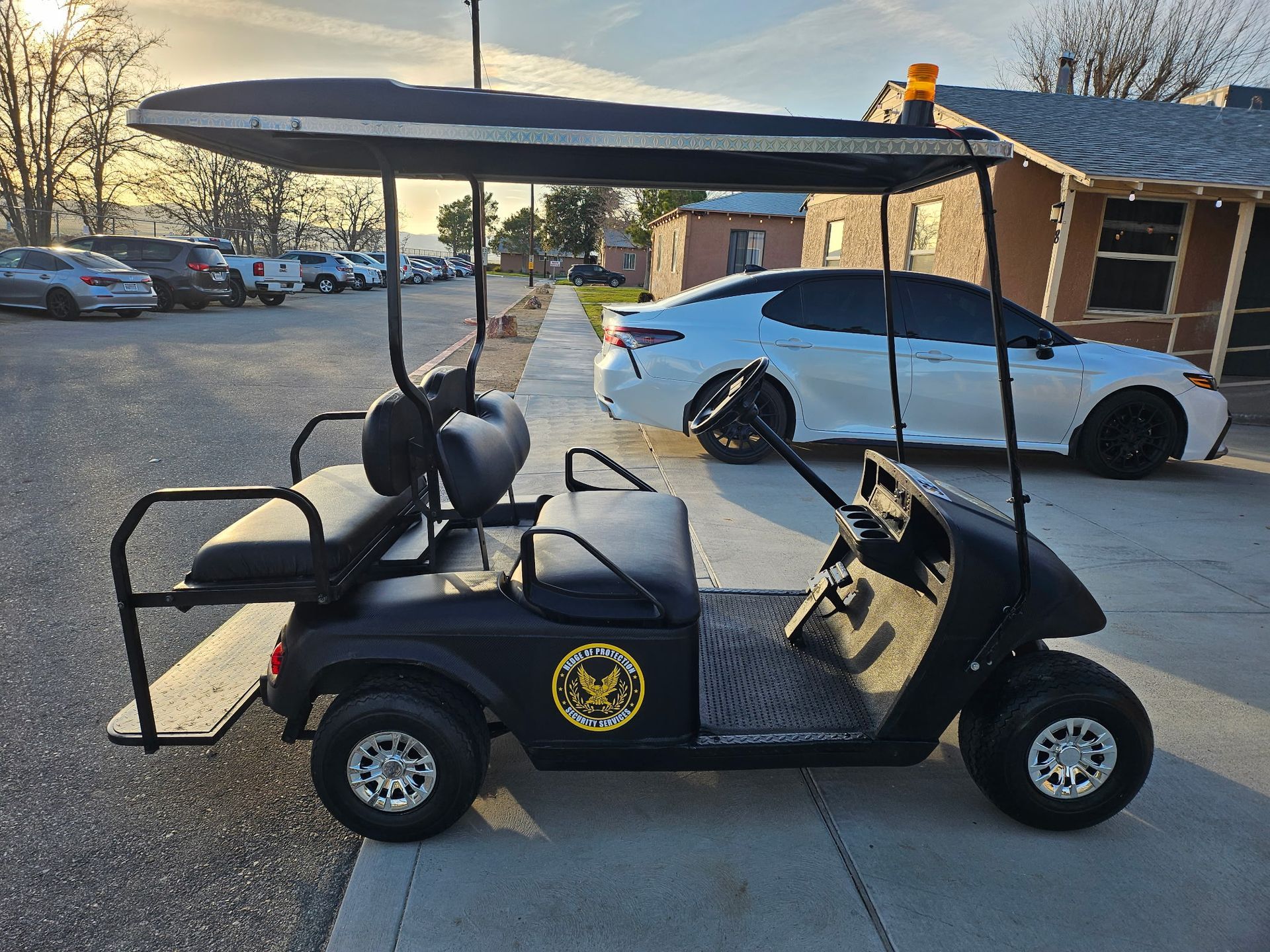 Black golf cart with yellow emblem parked under a canopy beside a white car at sunset.