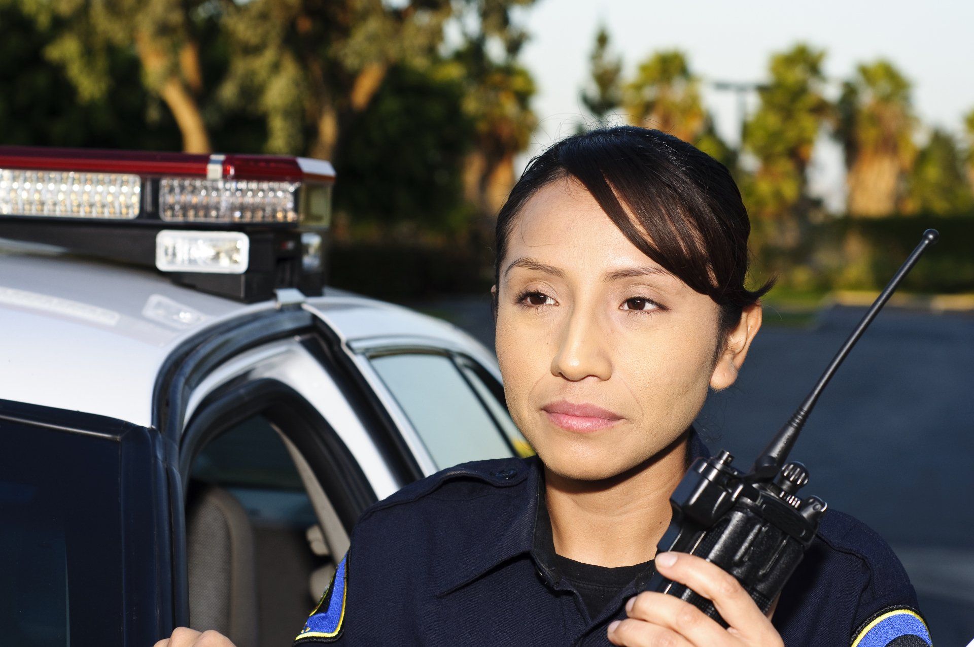 Police officer holding radio, standing near police car, looking thoughtful.