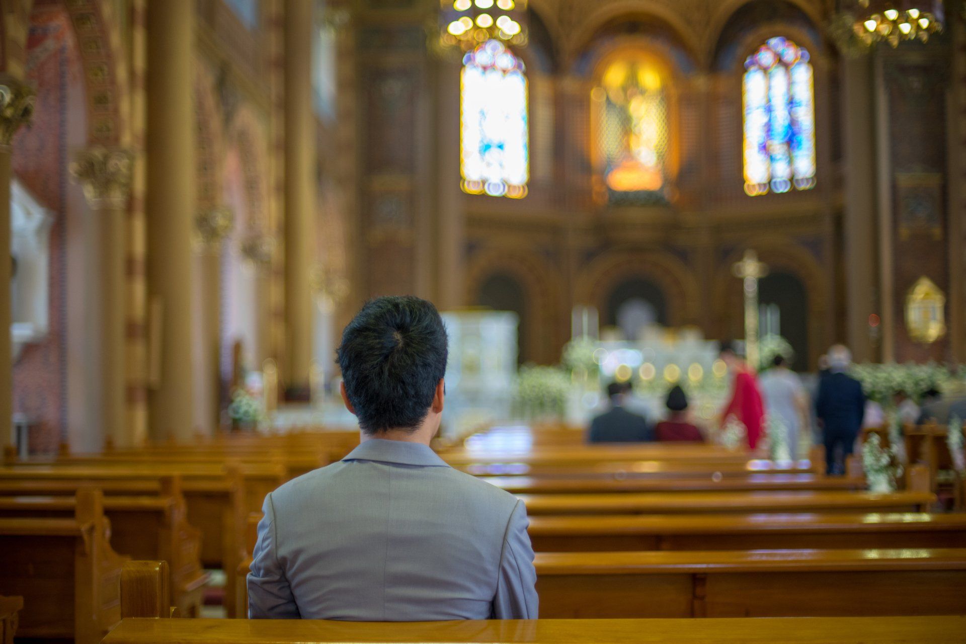 Person seated in church pew, facing altar. Stained glass windows and ornate architecture visible.