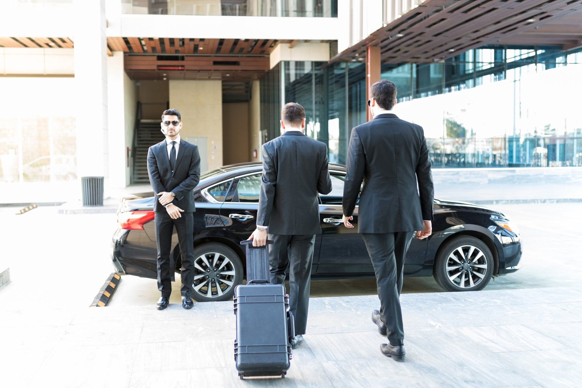 Three men in suits near a black car, one with luggage. A bodyguard stands watch near a building entrance.