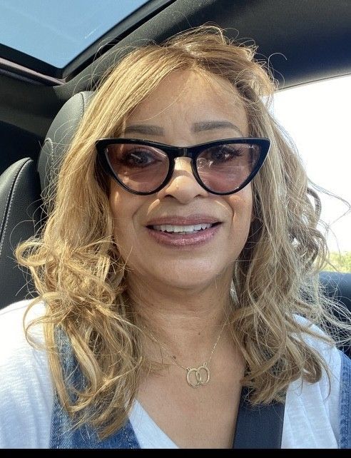 A woman wearing sunglasses and a necklace is smiling while sitting in a car.