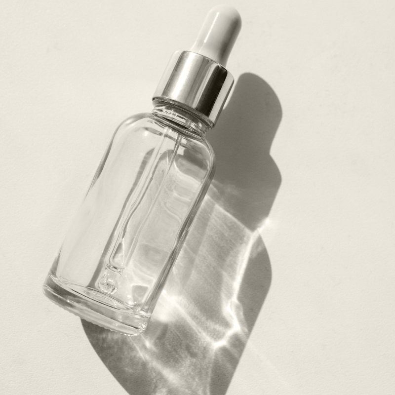 Clear glass dropper bottle with silver cap, lit by sunlight casting reflections on a white background