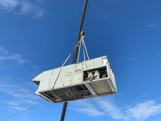 HVAC unit suspended by a crane against a bright blue sky.