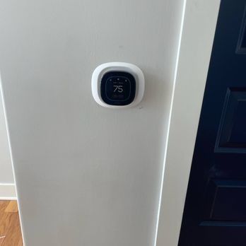 A white smart thermostat displaying 75 degrees is mounted on a white wall next to a dark blue door.