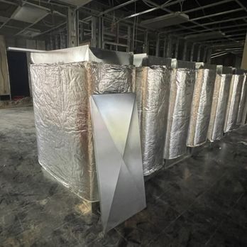 HVAC ductwork in construction. Silver insulated ducts line the room. A geometric metal shape stands in front.