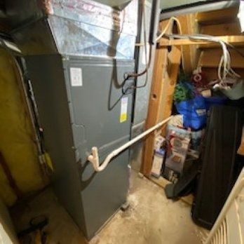Furnace in a cluttered basement corner. Gray metal with pipes and wires, beside storage items.