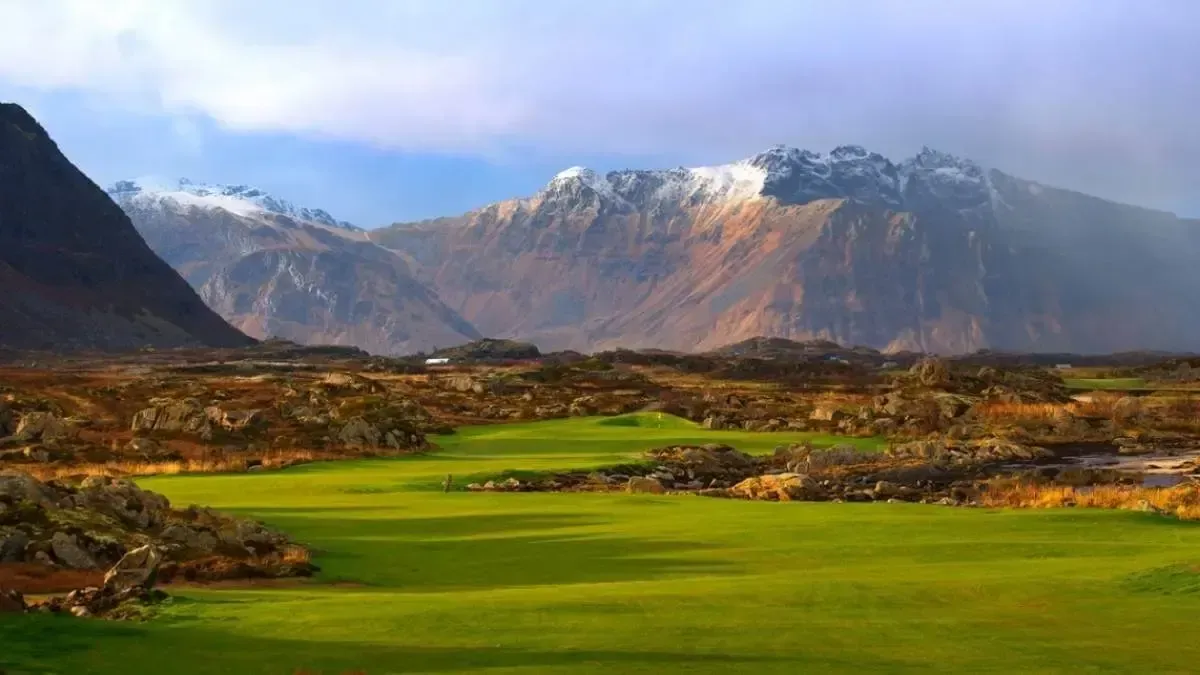 Green valley golf course beneath snow-capped mountains and a cloudy blue sky