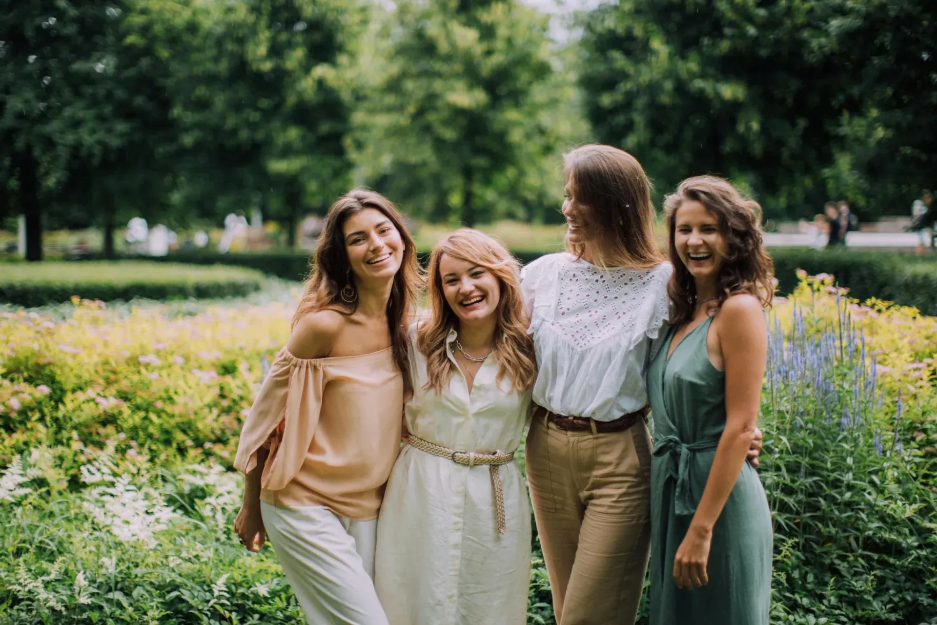 Four friends standing together in a lush green garden, smiling and posing in light summer outfits.