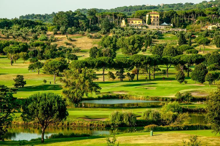 Lush green golf course with sand bunkers, water hazards, and distant buildings under trees