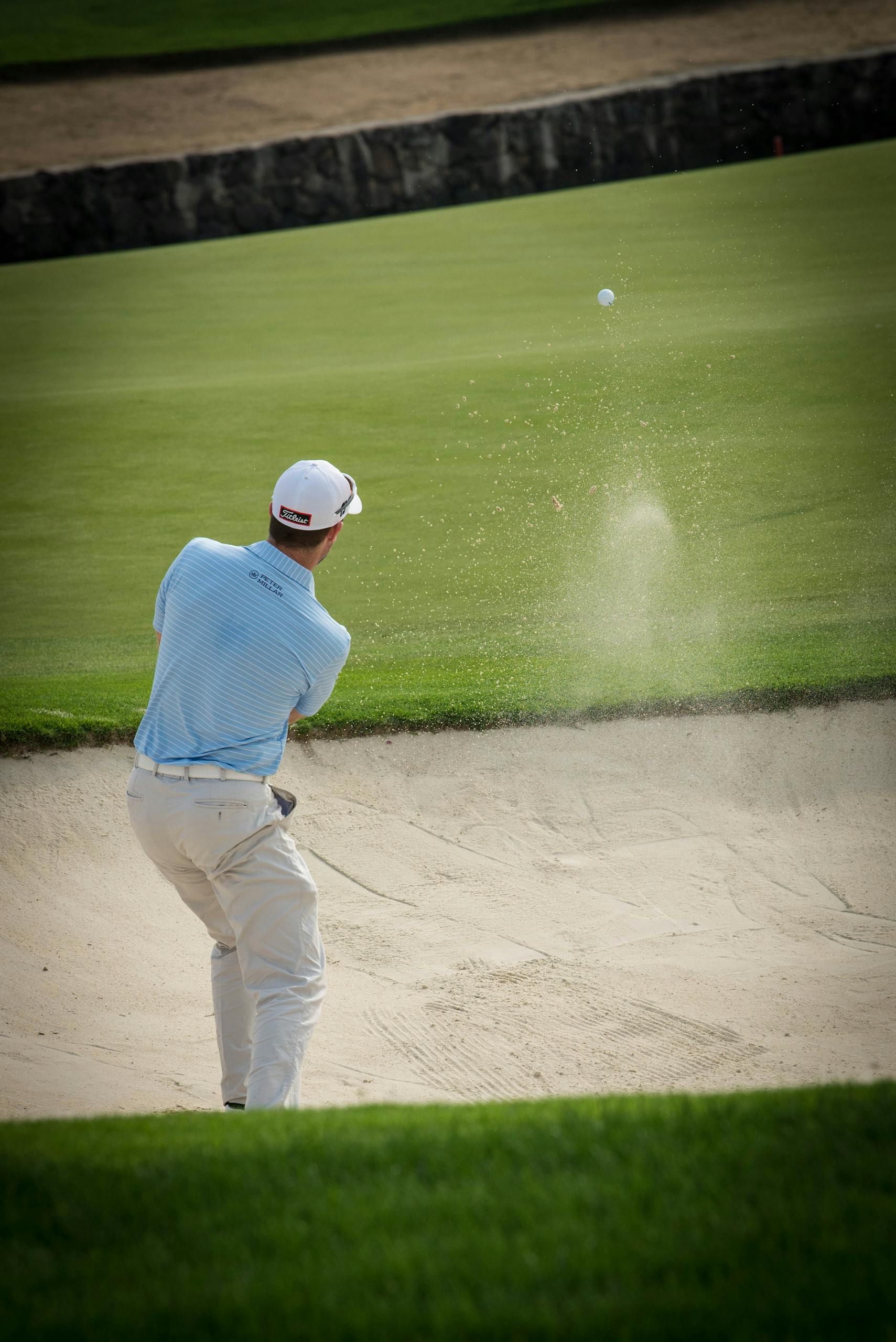 Golfer in light blue shirt swings from a sand bunker toward the green
