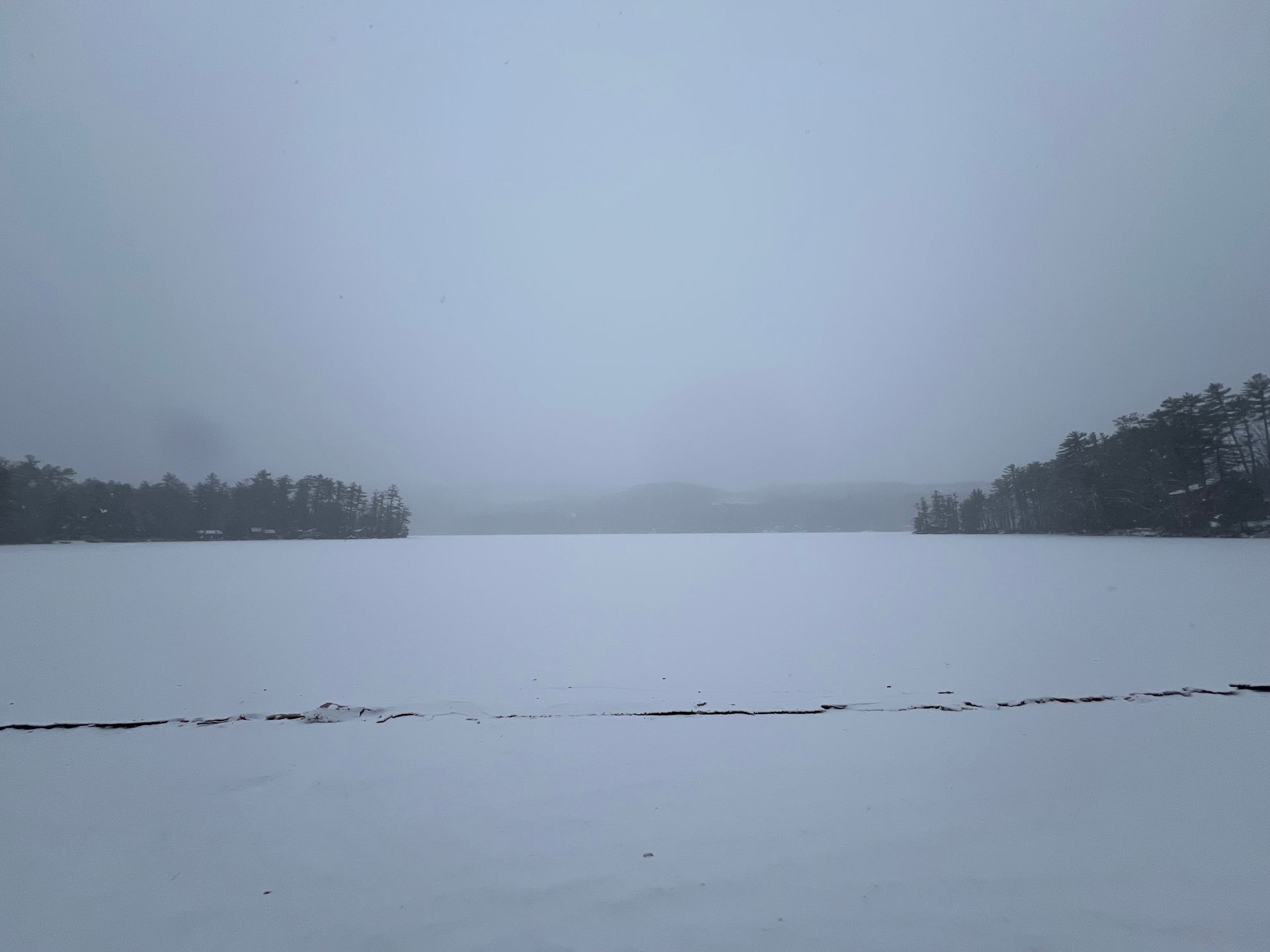 A snowy lake with trees in the background on a foggy day.