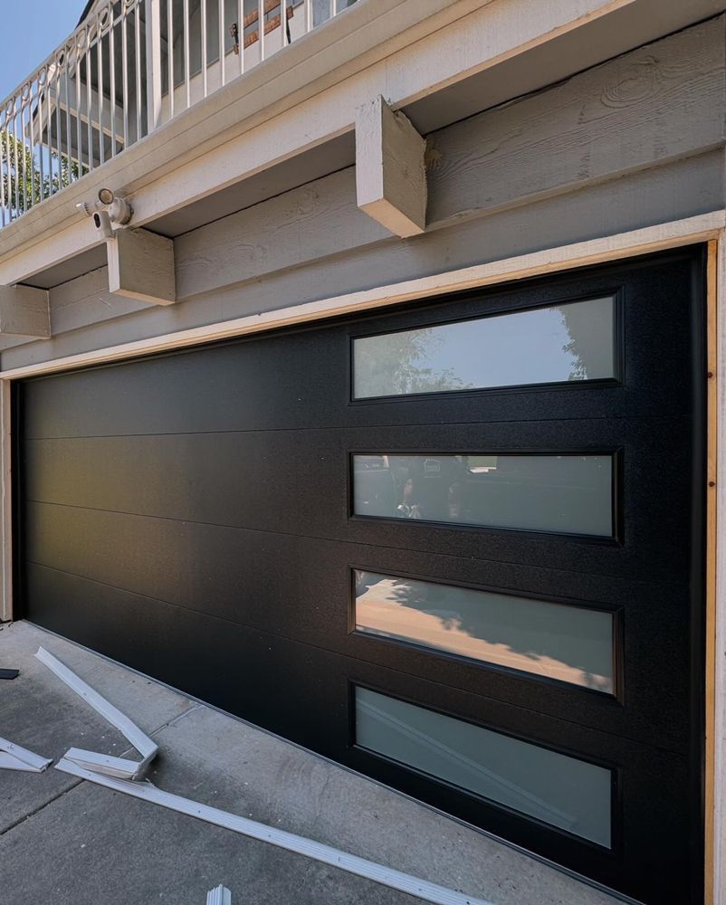 Black garage door with four horizontal windows under a balcony, beside a parking space marked by white lines