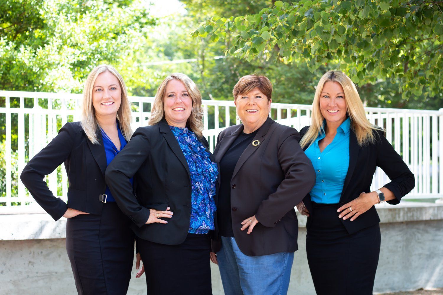 Four women in business attire posing outdoors, smiling. They stand in front of a white fence and greenery.