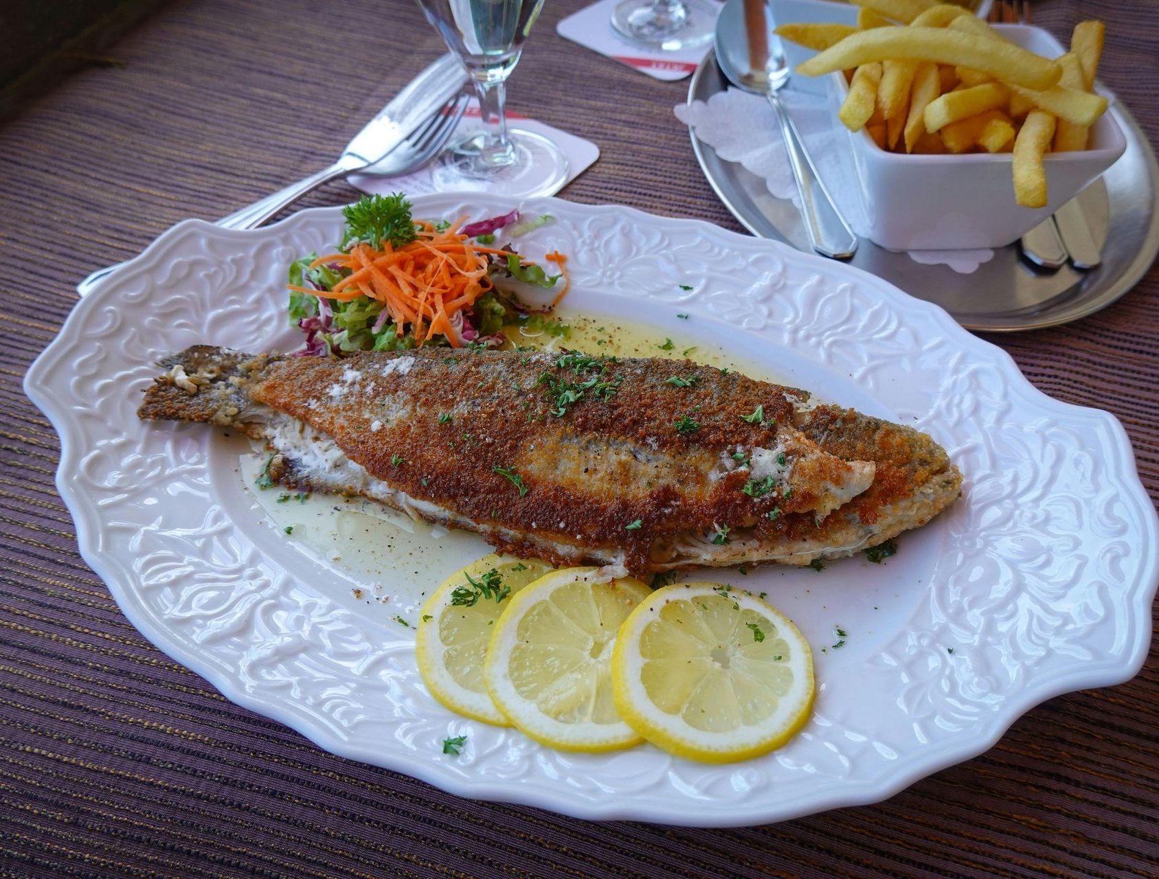 A plate of fried fish with lemon slices, salad, and a side of fries on a table in a restaurant.