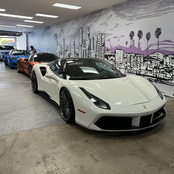 White sports car in a showroom, with several colorful cars lined up behind it.