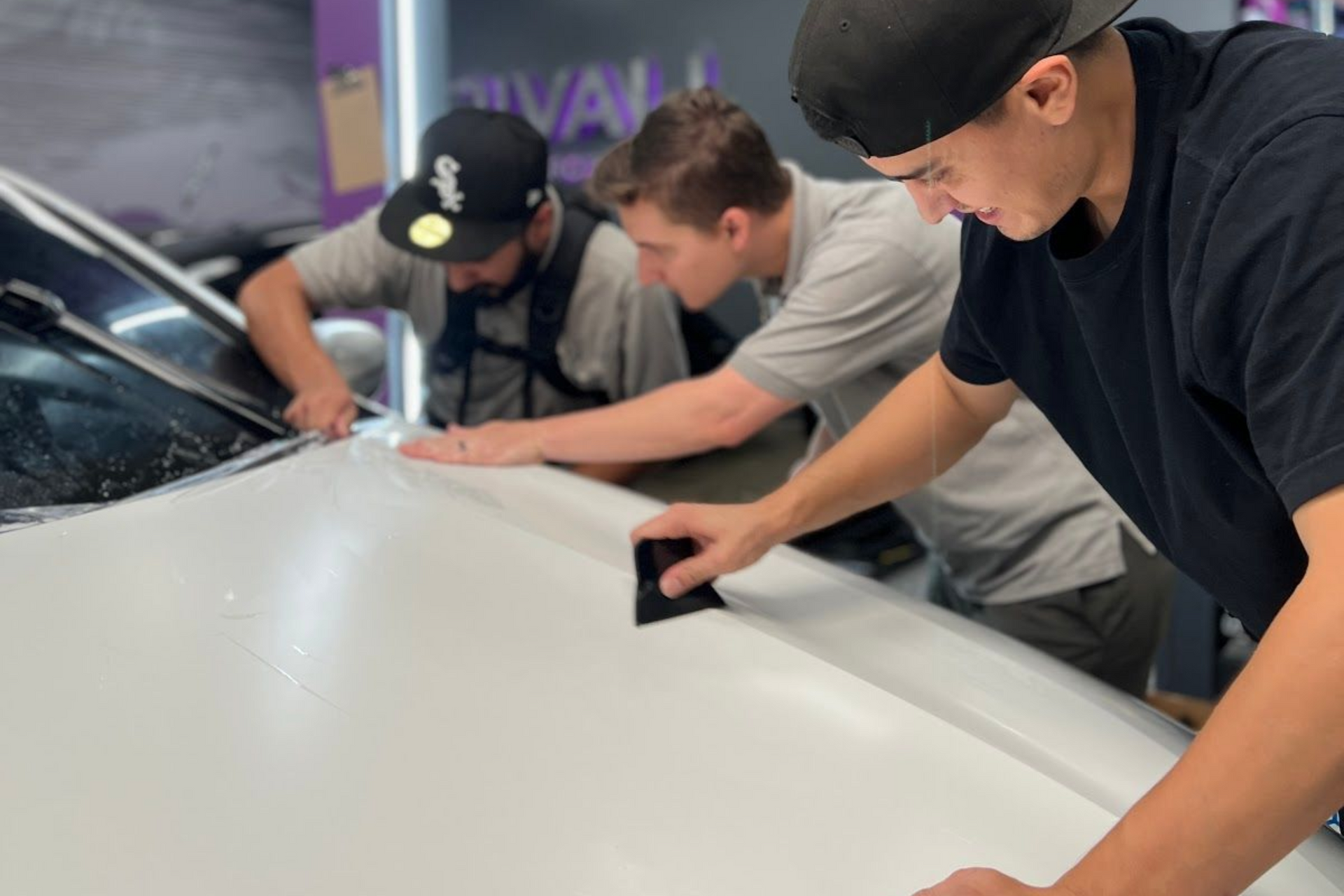 Three people working on a white car, applying a film. Shop interior with purple accents.
