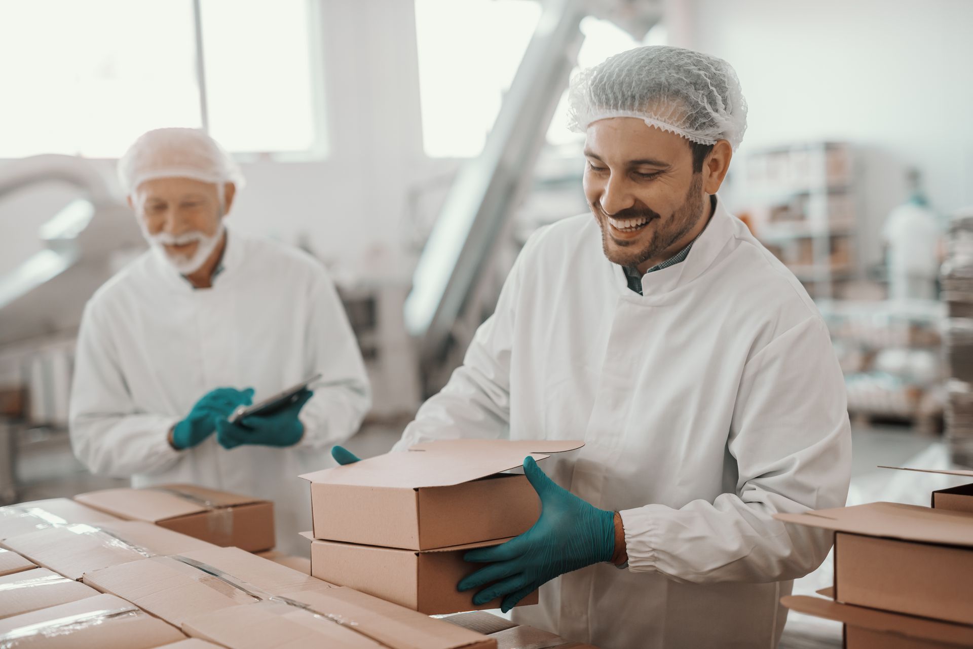 Two food workers in sanitary gear pack cardboard boxes in a bright, industrial production facility.