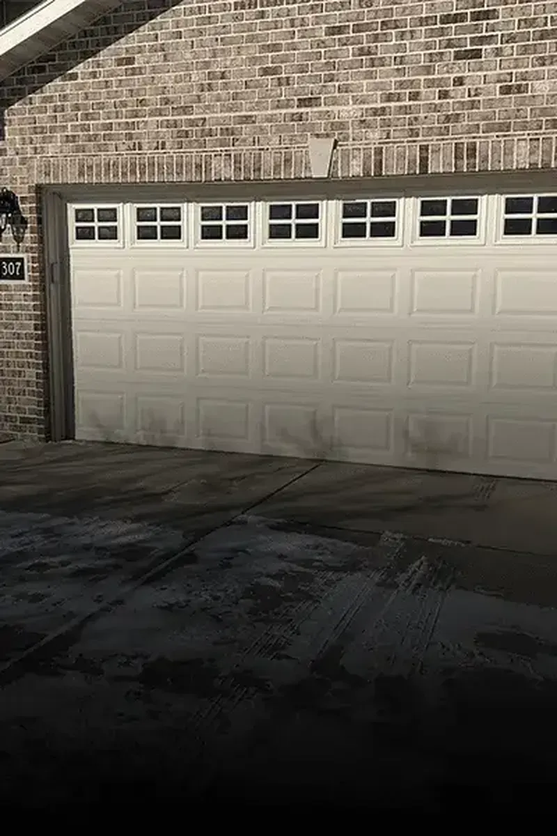 A white residential garage door with a row of small windows, set in a light-brown brick exterior wall.