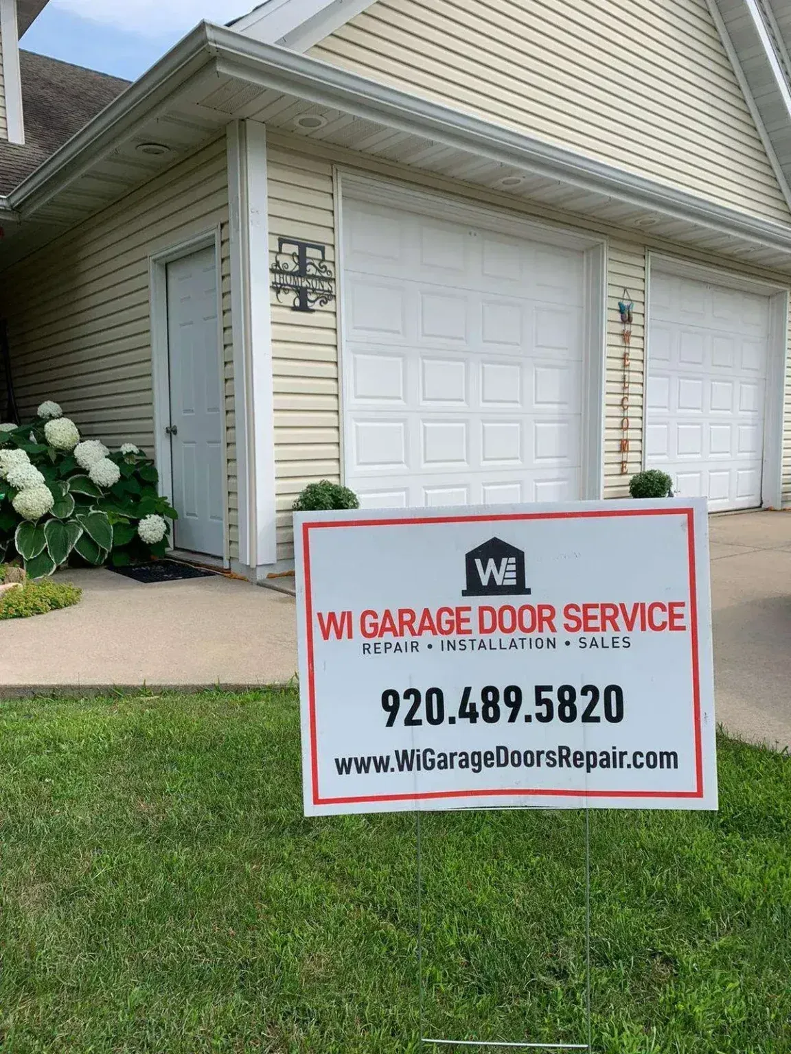 A sign for WI Garage Door Service in a yard in front of a house with two white garage doors and white siding.