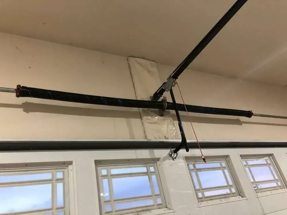 A black garage door torsion spring with a protective cover mounted horizontally above a white garage door with windows.