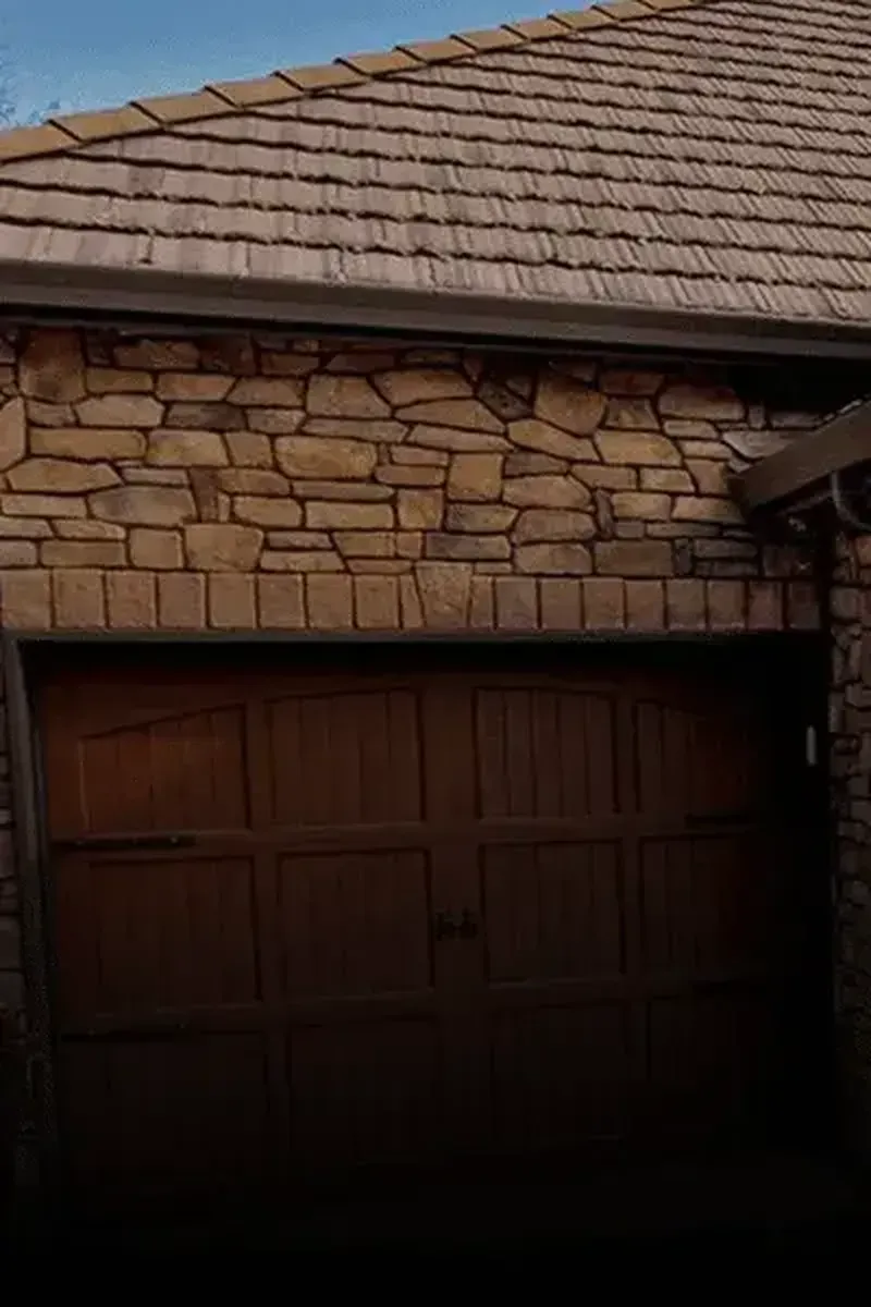A dark wood garage door sits below a horizontal row of stone bricks and an upper facade of irregular, tan-colored stones.