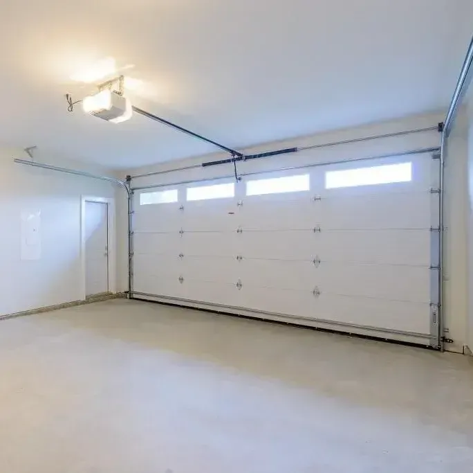 An empty garage with a large white sectional door, windows, and an overhead lighting system.