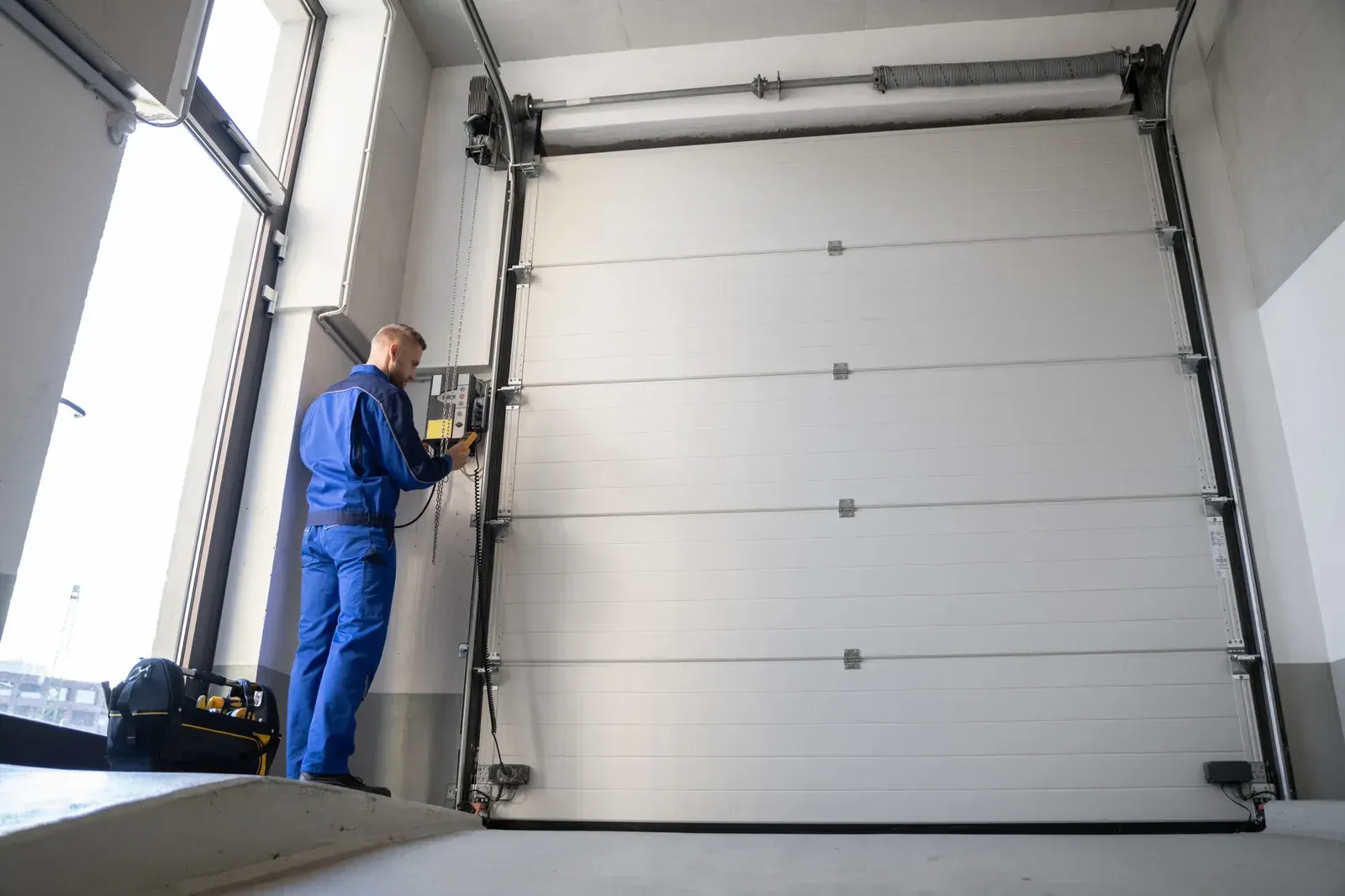 Technician in blue coveralls inspecting a large white garage door in a workshop