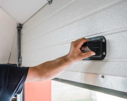 A person gripping a black handle on a white, horizontally paneled garage door.