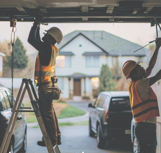 Two workers in hard hats installing a garage door, with a suburban house and truck in the background