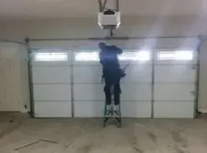 A person stands on a step ladder working on the ceiling-mounted garage door opener mechanism inside a garage.
