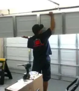 A technician in a dark uniform works to install or repair a white garage door inside a garage.