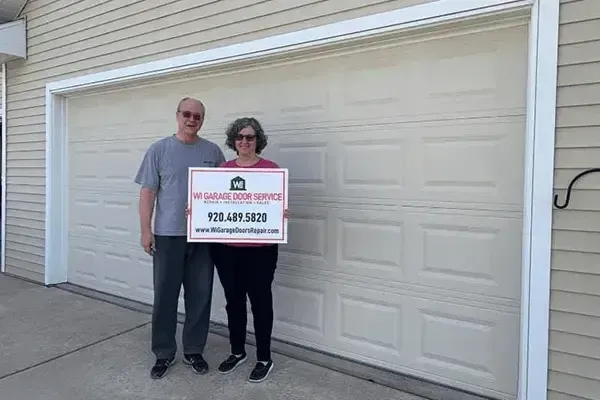 Two people stand in front of a new garage door holding a sign for a garage door repair service.