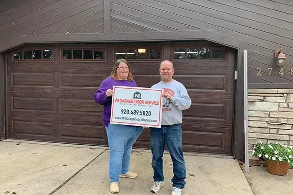 Two people stand in a driveway holding a sign for MR Garage Door Service in front of a house with a dark wood garage.
